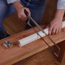 Man's hands cutting a white PVC pipe with a hacksaw.