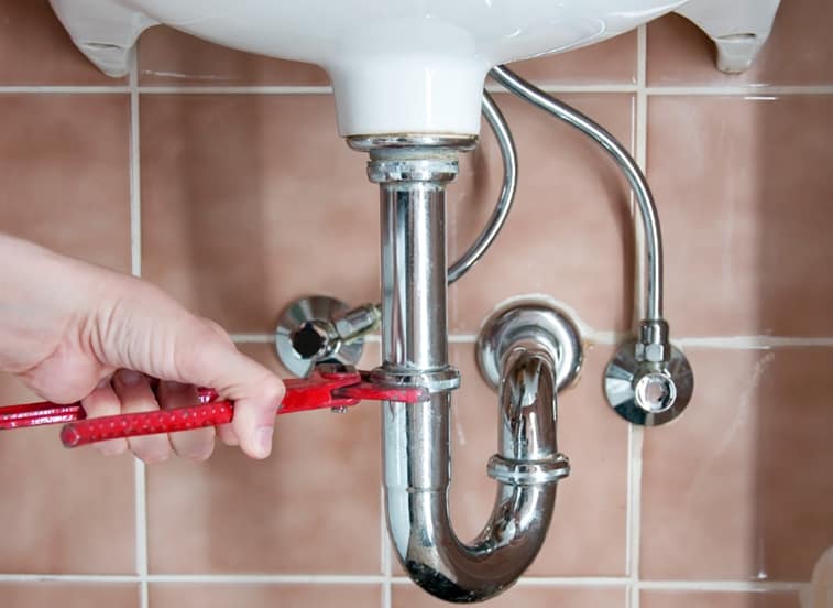 Man’s hand holding a slip-joint plier, loosening a sink drain’s coupling nut. 