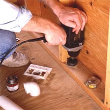 Man's hands using a power drill to bore a hole at a wall's bottom plate.