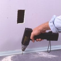 Man's hand drilling a small hole below a wall inlet opening.