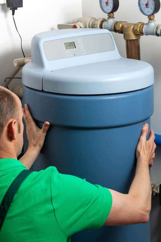 Man connecting a blue water softener to a house’s hot-water supply plumbing.