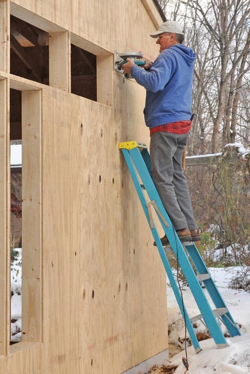 Man standing on a step-ladder, installing a house’s plywood exterior wall sheathing.