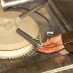 Man’s hands cleaning a dishwasher’s bottom spray hole with a stiff wire.