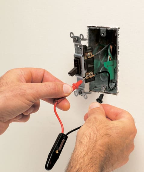 Man’s hands holding a neon tester’s insulated parts including a red probe tip touching a brass terminal screw and a black probe tip meeting a metal box screw hole.