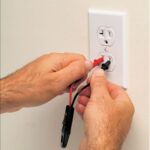 Man’s hands holding a neon tester’s insulated parts with red and black probe needle-tips inserted inside the slots of a T-shaped power outlet.