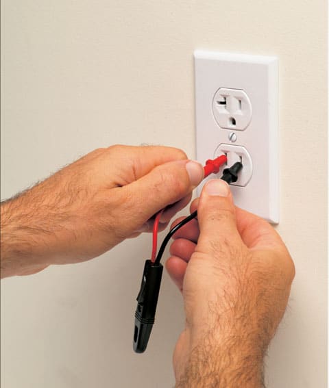 Man’s hands holding a neon tester’s insulated parts with red and black probe needle-tips inserted inside the slots of a T-shaped power outlet.