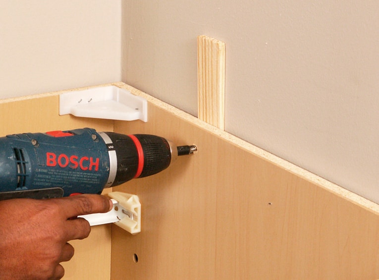 A hand using a screw gun to attache the backs of cabinets and shims into wall studs.