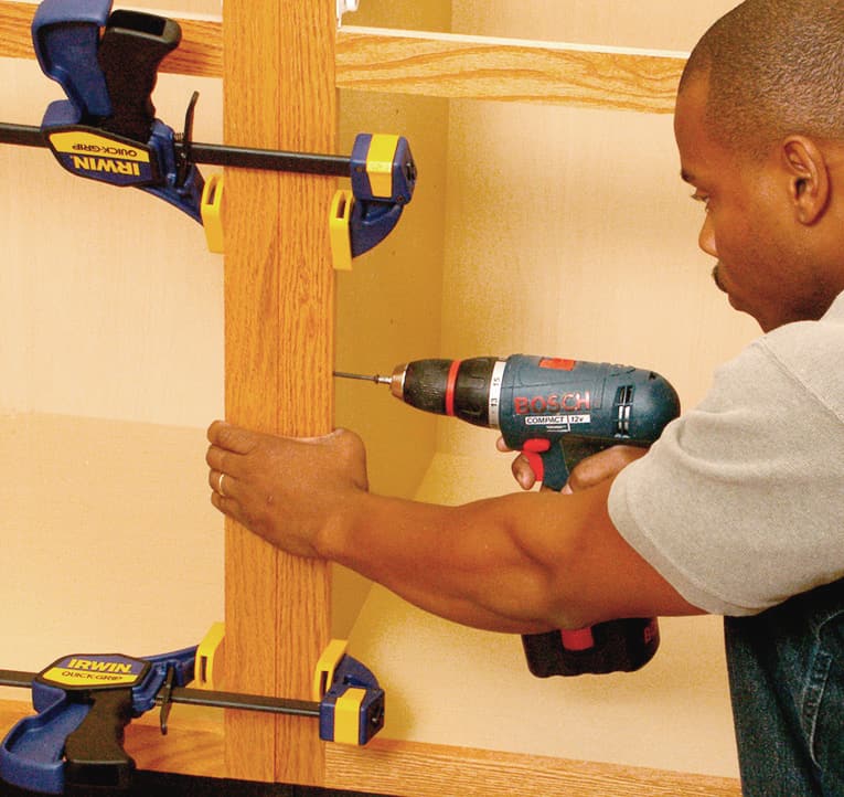 A man clamping together adjoining cabinets, and screwing through the edge of one face frame into the other.