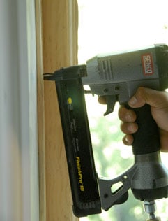 Man's hand using a pneumatic nailer on a window molding.