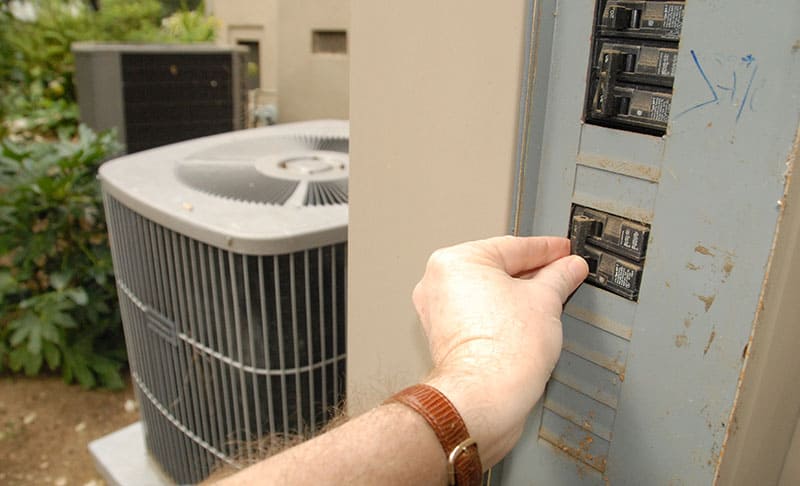 Hand turning off an outdoor circuit breaker next to a central air conditioner compressor. 