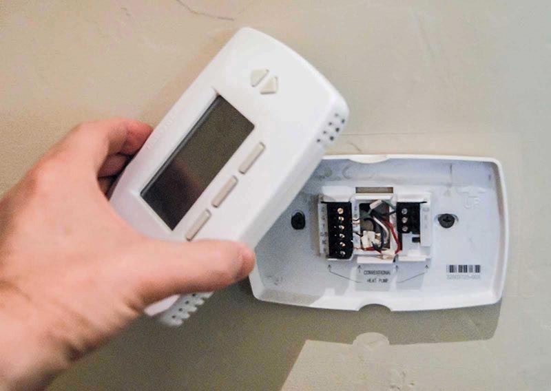 Man’s hand holding an electronic thermostat cover exposing colored internal wires.