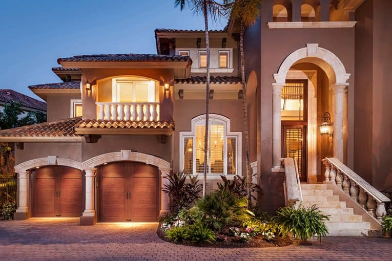Two-story Mediterranean house’s front yard including an outdoor landscape and a carriage-style, wooden double door garage.