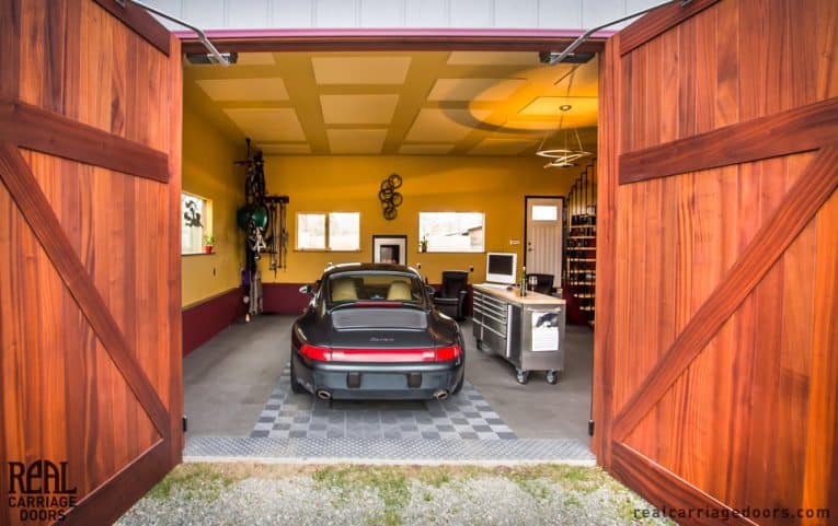 Open solid-wood doors in a swing-out carriage garage including a sports car, a stainless metal tool cabinet with a monitor on top, an armchair, a pendant ceiling light, and a spiral staircase.