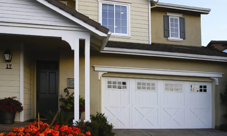 Two-story bungalow house’s front yard including some flowers and a large carriage-style, white wooden door garage.