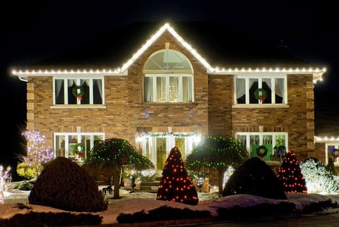 Two story brick house at night, with Christmas lights on the roofline and in the garden.