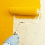 Man’s hand painting a wall with yellow using a roller.