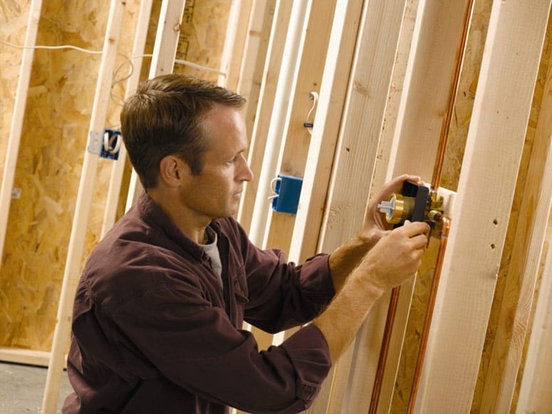 Man installing a shower valve between a house’s interior wall studs.