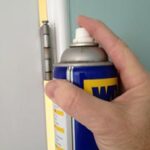 Man’s hand spraying a can of WD-40 lubricant on a door hinge.