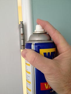 Man’s hand spraying a can of WD-40 lubricant on a door hinge.