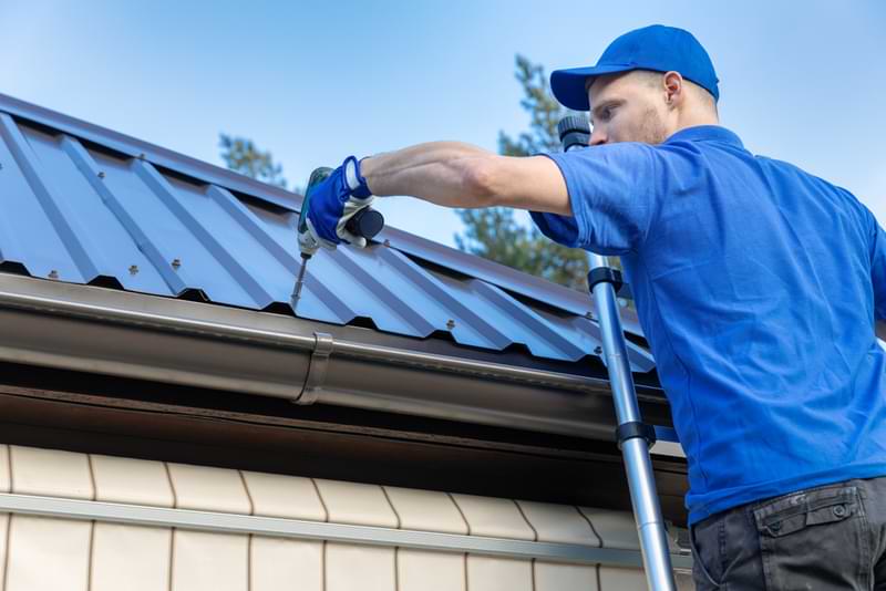 A roof installer on a ladder screwing in new metal roofing panels. 