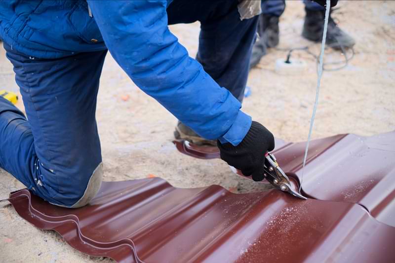 A worker cutting a piece of brown metal roofing with hand sheers. 