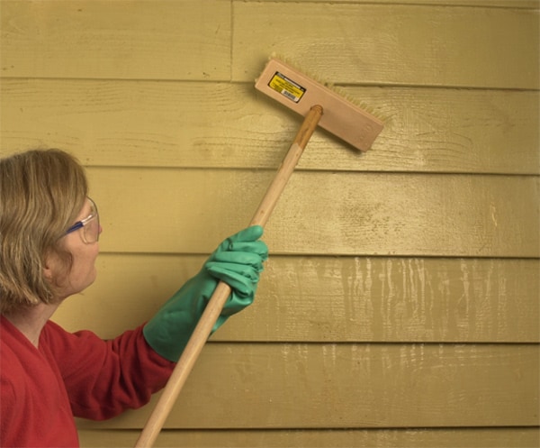 A man using a wooden brush with long handle to scrub house siding.