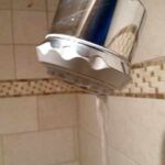 Water leaking out of a chrome showerhead in a tiled shower.