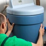 Man connecting a blue water softener to a house’s hot-water supply plumbing.