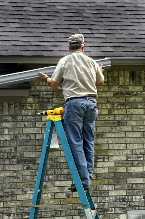 Man standing on a step ladder, installing a house’s rain gutter. 