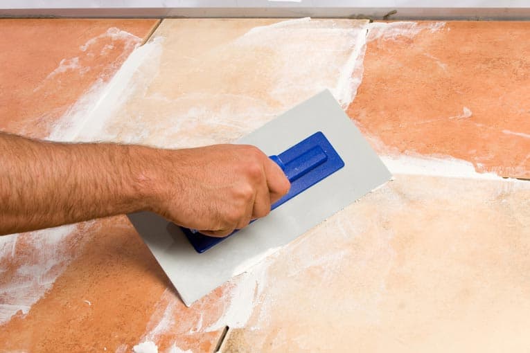 Man's hand applying grout to joints between tiles, using a grout float.