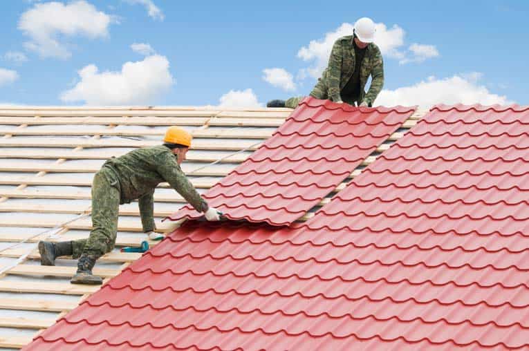 Two men installing a red metal roofing over wooden battens.