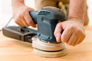 Man's hands sanding a wooden surface with an orbital sander.