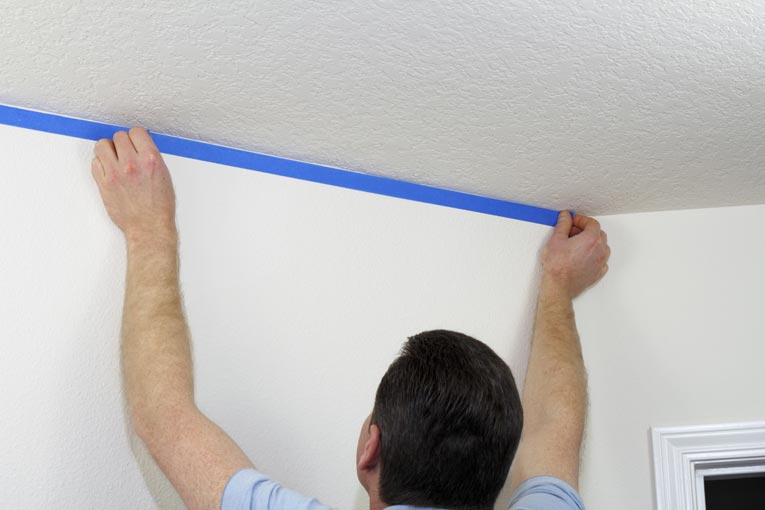Man placing a strip of blue painter's tape along a wall's top most corner.