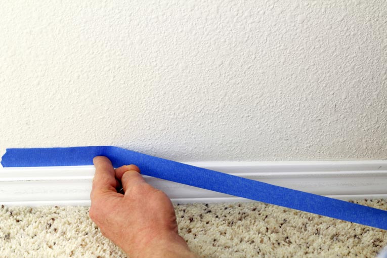 Man's hand masking a wall’s base, next to a trim molding.