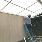 A man standing on a step ladder, anchoring a steel and polycarbonate patio roof.