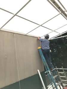 A man standing on a step ladder, anchoring a steel and polycarbonate patio roof.