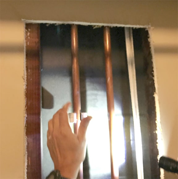 Man’s hand using a tubing cutter on pipe, behind a cut-out wall.