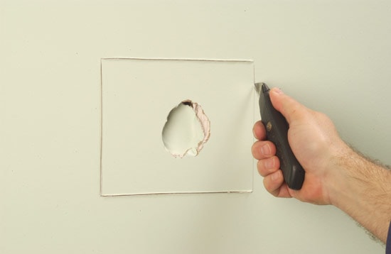 Hand using utility knife to make rectangular cutout around drywall hole