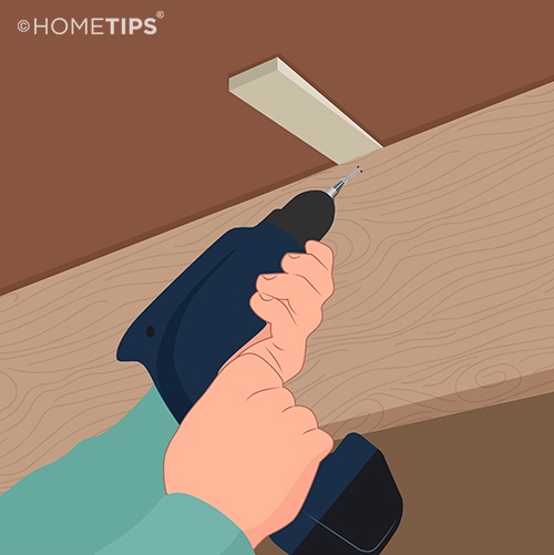 Hands using a power drill to drive a screw through a joist and into a wood shim beneath the floor.