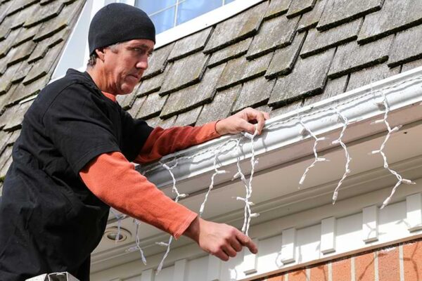 Man hanging icicle lights on the gutter of a home with a wood shingle roof.